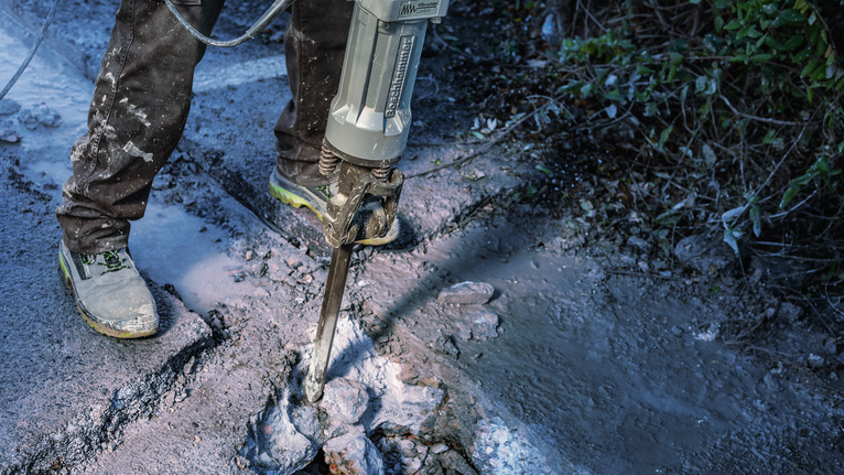 Worker wearing safety equipment breaks concrete with a demolition hammer on a wet surface.