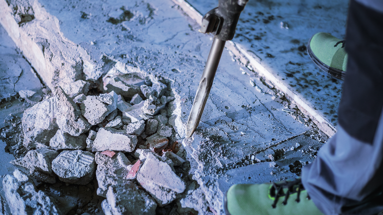 Person wearing safety equipment breaks concrete with a demolition tool.