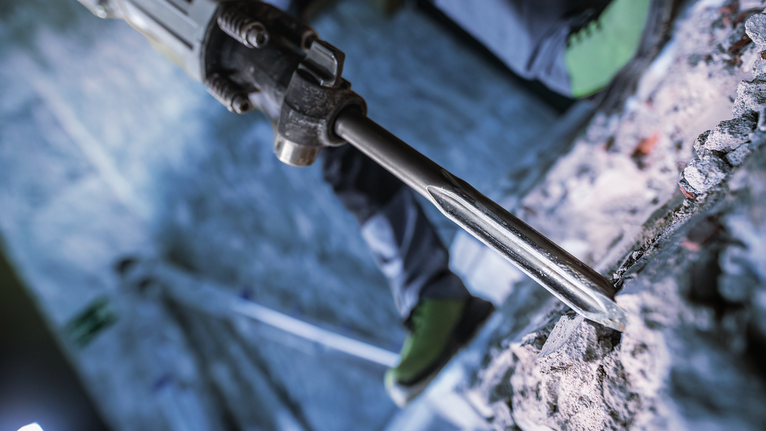 Person wearing safety equipment operates a demolition hammer breaking concrete.
