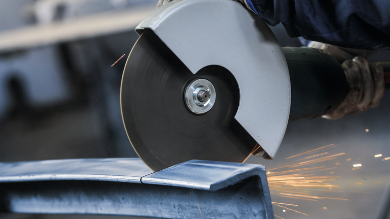 A person wearing safety equipment cuts metal with a large angle grinder.