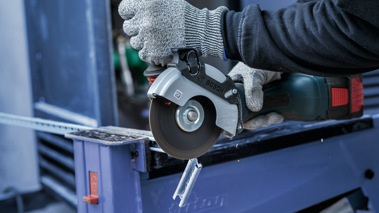 Person wearing safety equipment uses an angle grinder to cut a metal bar.