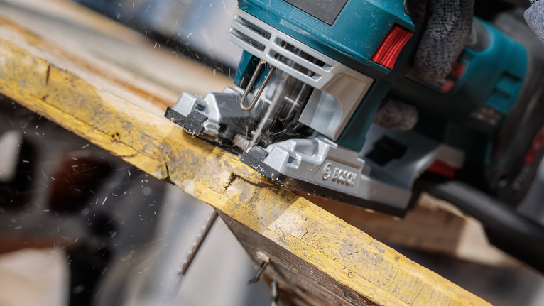 Worker wearing safety equipment uses a jigsaw to cut through a wooden plank.