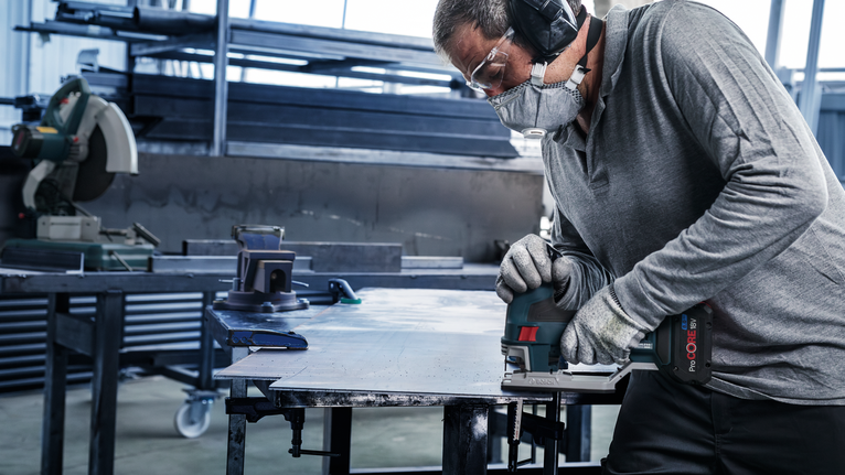 Worker wearing safety equipment uses a cordless jigsaw to cut a metal sheet.