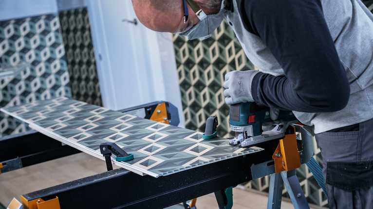 A worker wearing safety equipment uses a jigsaw to cut patterned tile.