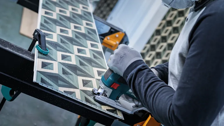 A worker wearing safety equipment cuts patterned board with a handheld saw.