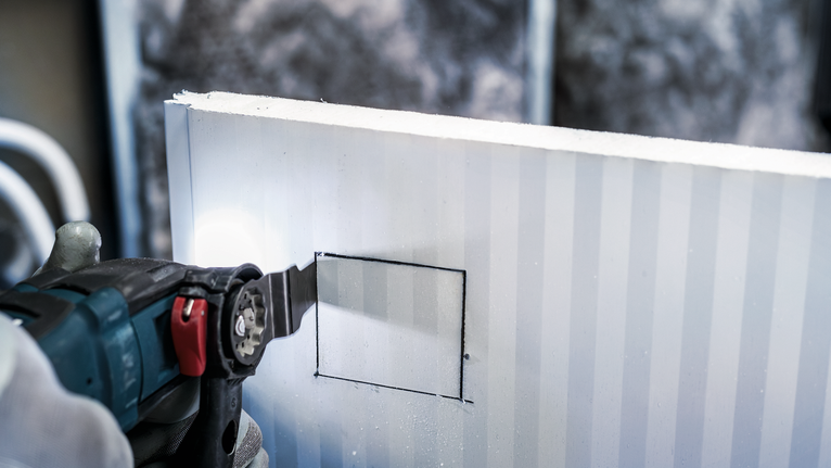 Person wearing safety equipment cutting a square opening in a panel with a power tool.