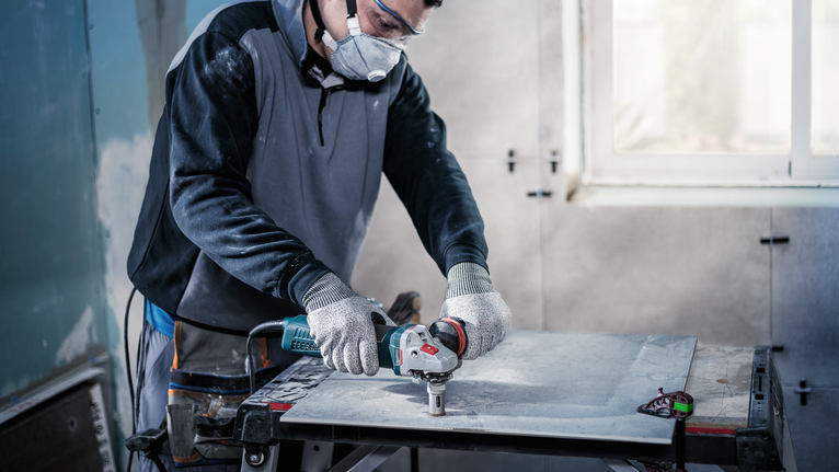 Person wearing safety equipment uses a power tool to cut a large tile indoors.