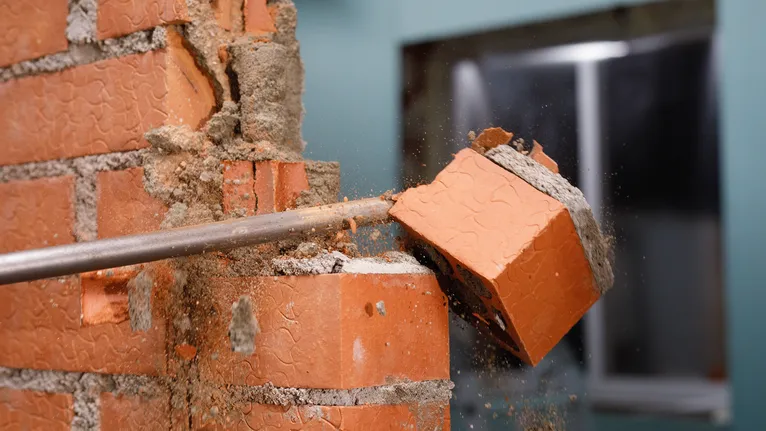 A chisel breaks a brick from a wall, sending dust and debris flying.