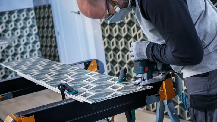 Person wearing safety equipment cuts patterned tile with a jigsaw on a workbench.
