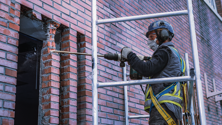 Person wearing safety equipment drills into a brick wall beside scaffolding.