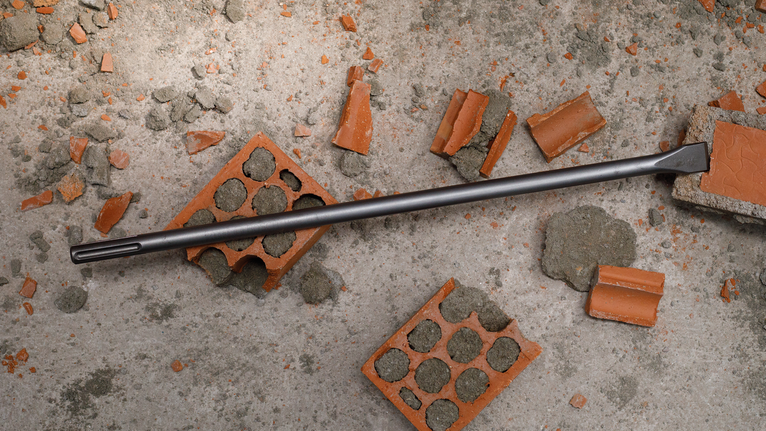 Chisel drill bit lying on concrete surface surrounded by broken bricks and debris.