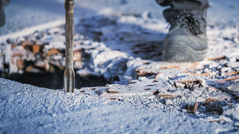 Person wearing safety equipment uses a demolition tool to break concrete flooring.