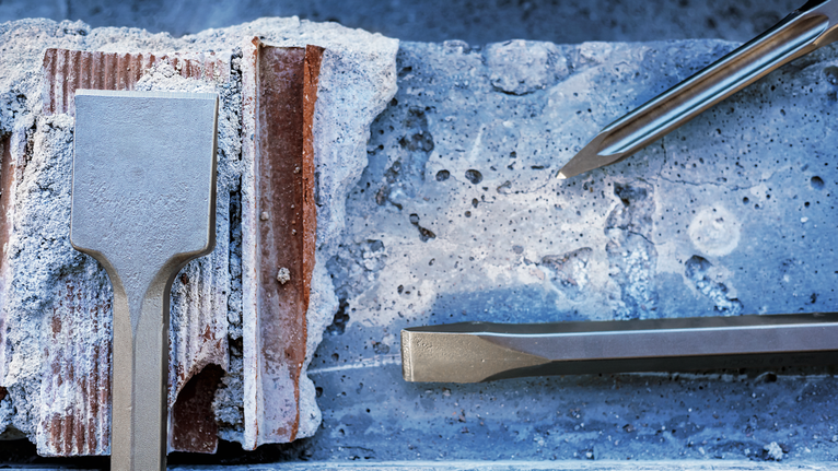 Three metal chisel bits rest on rough concrete and brick rubble.