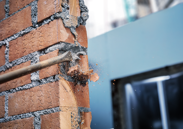 Chisel tool breaking through a brick wall, scattering debris.
