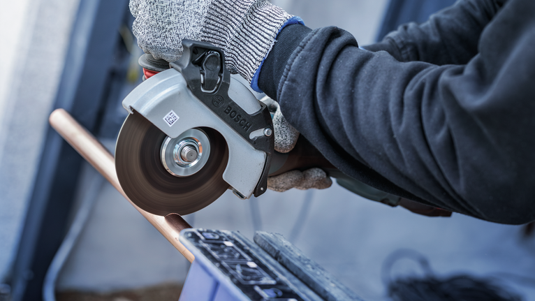Person wearing safety equipment uses an angle grinder to cut a metal pipe.
