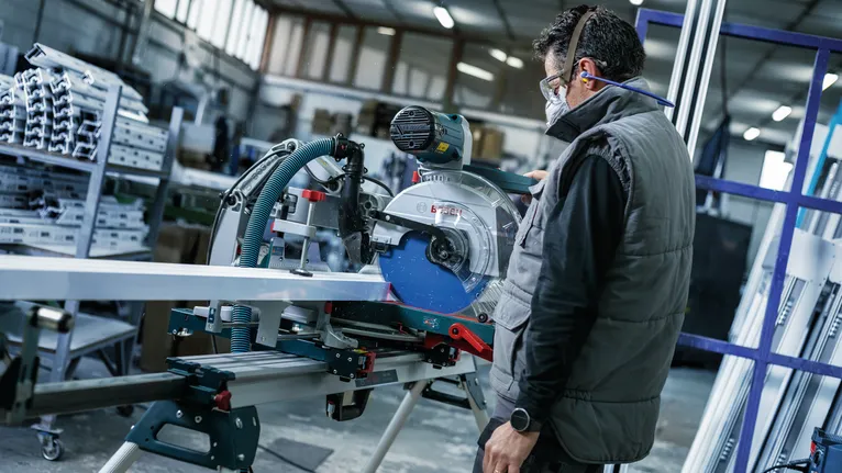 Person wearing safety equipment cuts metal pipe with a sliding miter saw in a workshop.