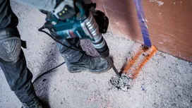 Person wearing safety equipment drills into concrete on a marked construction site floor.