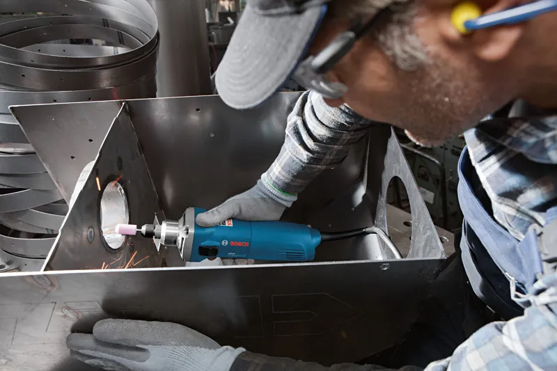 A person wearing safety equipment uses a straight grinder tool on a metal sheet.