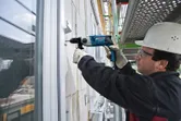 Une personne portant un équipement de sécurité utilise une perceuse à percussion pour enfoncer une vis dans un mur.