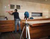 Une personne marque des carreaux muraux dans une cuisine à l'aide d'un outil de nivellement laser sur un trépied.