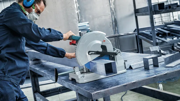 Une personne portant un équipement de sécurité coupe une poutre métallique avec une scie circulaire dans un atelier.