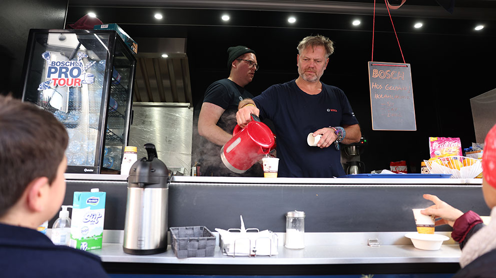 Bosch standında bir adam, fincana sıcak içecek döküyor, arka planda diğer adam var.