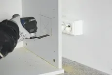 Güvenlik ekipmanı giyen bir kişi, elektrikli bir alet kullanarak ahşap bir panelde kare şeklinde bir açıklık kesiyor.