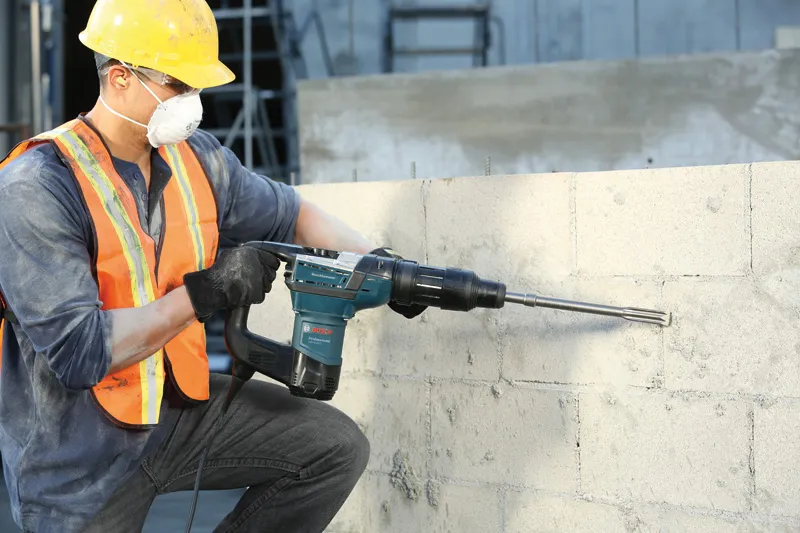 Güvenlik ekipmanı takan bir kişi beton duvara matkapla delme işlemi yapıyor.