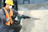 Güvenlik ekipmanı takan bir kişi beton duvara matkapla delme işlemi yapıyor.