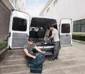 Açık bir kamyonetin yanında duran iki işçi aletleri düzenliyor ve kablosuz elektrikli süpürge kullanıyor.