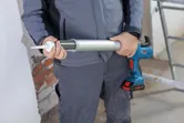 Bir kişi tuğla duvarın yakınında sızdırmazlık maddesi uygulamak için kablosuz bir macun tabancası tutuyor.
