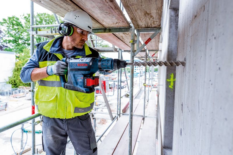 Bir inşaat sahasında güvenlik ekipmanı takan bir kişi beton duvara matkapla delik açıyor.