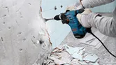 Güvenlik ekipmanı takan bir kişi, duvardaki fayansları sökmek için döner çekiç kullanıyor.