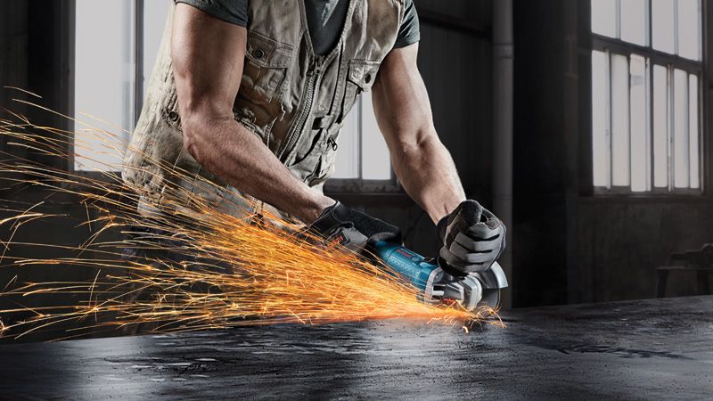 Güvenlik ekipmanı giyen bir kişi, açılı taşlama makinesi kullanarak metal taşlıyor, kıvılcımlar saçılıyor.