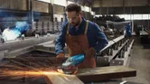 Fabrikada güvenlik ekipmanı takan bir kişi metal öğütüyor, kıvılcım çıkıyor.