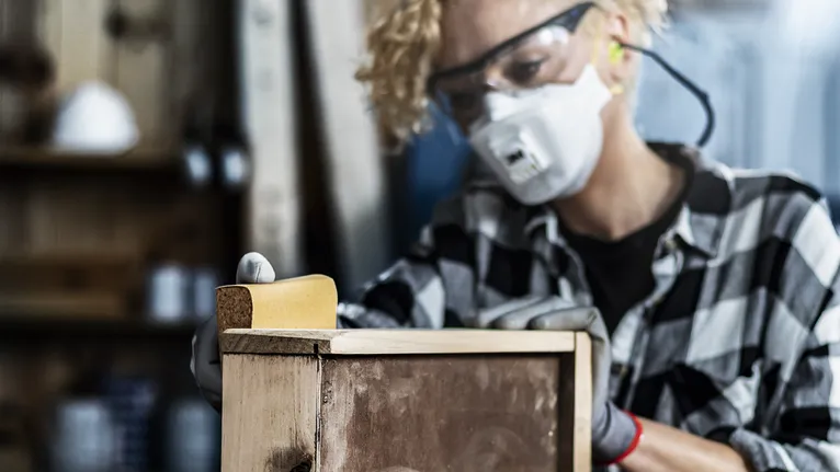 Bir atölyede güvenlik ekipmanı takan bir kişi tahta bir kutuyu zımparalıyor.