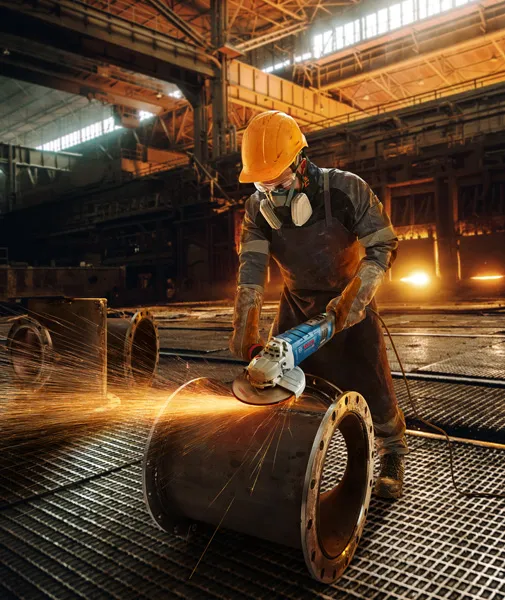 Güvenlik ekipmanı takan bir kişi büyük bir metal boruyu aşındırıyor ve kıvılcımlar çıkıyor.