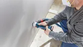 Güvenlik ekipmanı takan bir kişi, kablosuz alçıpan frezesi kullanarak alçıpanda delik açıyor.