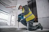 Güvenlik ekipmanı takan bir kişi, bir inşaat sahasında betonu kırmak için darbeli matkap kullanıyor.