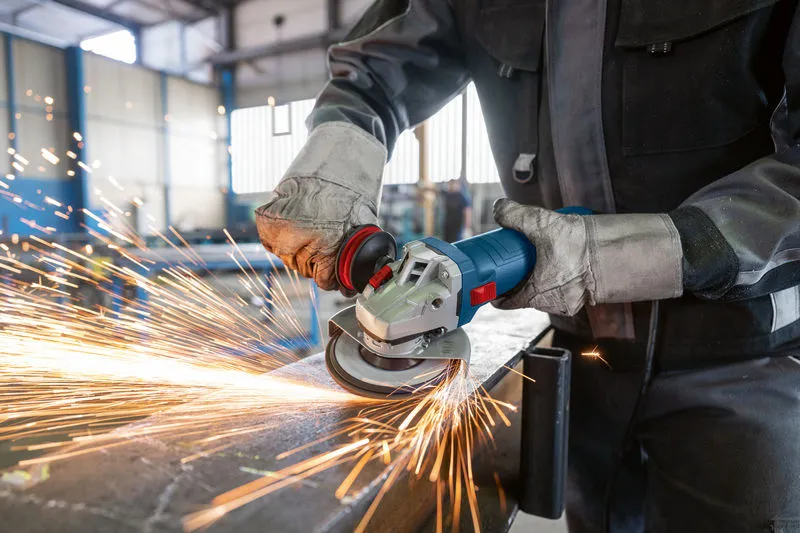 Güvenlik ekipmanı takan bir kişi açılı taşlama makinesi kullanarak kıvılcımlar saçarak metal öğütüyor.