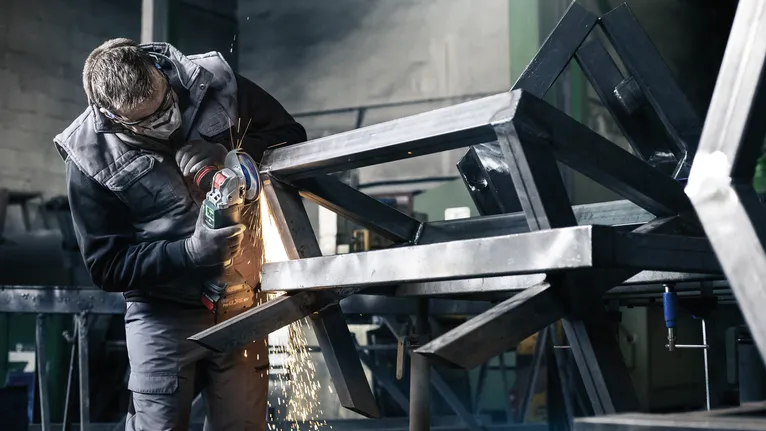Güvenlik ekipmanı takan bir kişi metal bir çerçeveyi aşındırıyor ve kıvılcımlar çıkıyor.