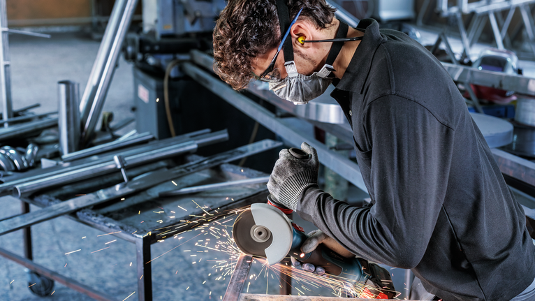 Güvenlik ekipmanı takan bir kişi açılı taşlama makinesi kullanarak metal keserken kıvılcımlar çıkarıyor.