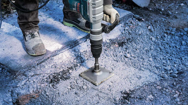 Güvenlik ekipmanı takan bir kişi, dışarıda beton kırmak için elektrikli matkap kullanıyor.