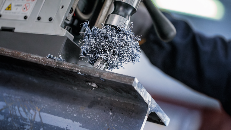 Güvenlik ekipmanı takan bir kişi, metal bir kirişe matkapla delik açarak metal talaşı oluşturuyor.