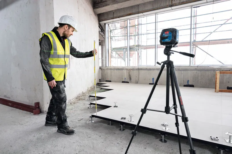 Güvenlik ekipmanı takan bir kişi, bir tripod üzerindeki lazer tesviye aletini kullanarak zemini tesviye ediyor.