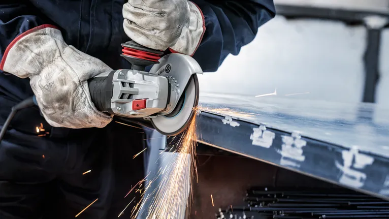 Güvenlik ekipmanı takan bir kişi açılı taşlama makinesiyle metal bir sacın kenarını taşlar.