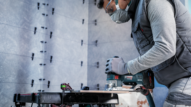 Güvenlik ekipmanı takan bir kişi, bir çalışma tezgahında odun kesmek için elektrikli testere kullanıyor.