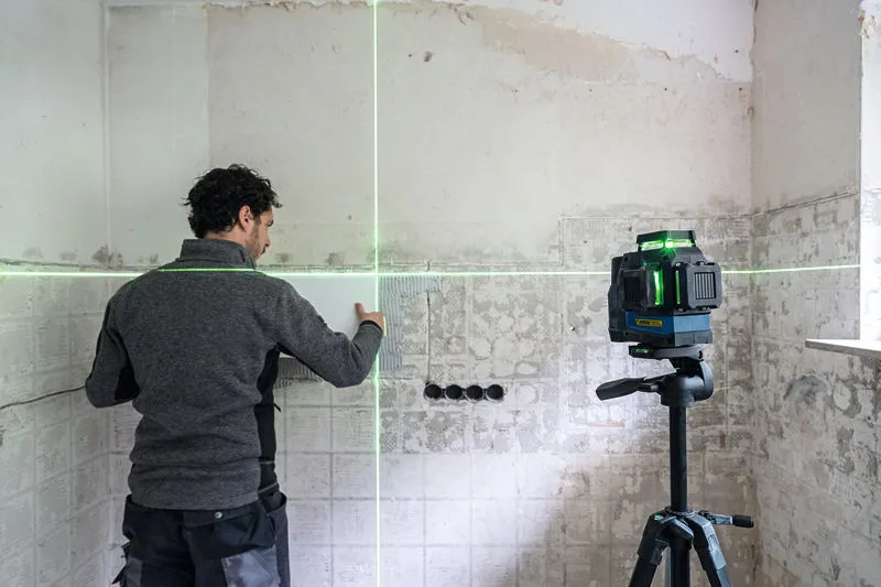 Bir kişi, bir tripod üzerindeki yeşil lazer tesviye aletini kullanarak duvardaki fayansları hizalıyor.