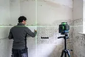 Bir kişi, bir tripod üzerindeki yeşil lazer tesviye aletini kullanarak duvardaki fayansları hizalıyor.