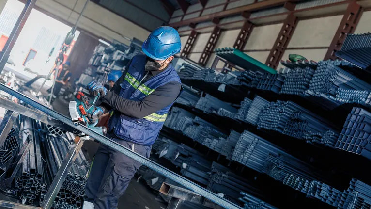 Güvenlik ekipmanı giyen bir kişi, endüstriyel bir depoda metal kesmek için elektrikli testere kullanıyor.